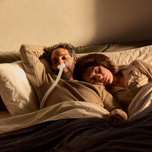 Load image into Gallery viewer, Two people lying in bed together with a blanket and pillow, one person using an oxygen mask.