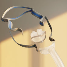 Load image into Gallery viewer, A sleep apnea mask with a clear nose cup and transparent tubing, attached to a black and blue headband, shown against a warm beige background with soft sunlight shadows.