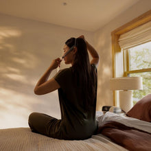 Load image into Gallery viewer, A woman sits on a bed in a sunlit bedroom, adjusting her hair with a hairbrush. The room has warm sunlight streaming across the wall, a window with a wooden frame, and a bedside lamp.