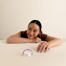 Load image into Gallery viewer, A woman with dark hair tied back leans forward on a cream-colored table, smiling at a small purple and white mushroom-shaped object placed in front of her.
