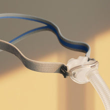 Load image into Gallery viewer, A close-up of a sleep apnea mask with a gray and blue head strap connected to a clear mouthpiece and transparent breathing tube, set against a warm beige background with soft sunlight.

