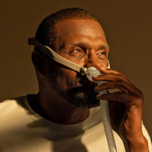 Load image into Gallery viewer, A man wearing a white long-sleeve shirt and holding a white nasal oxygen mask with a clear tube against a warm brown background.
